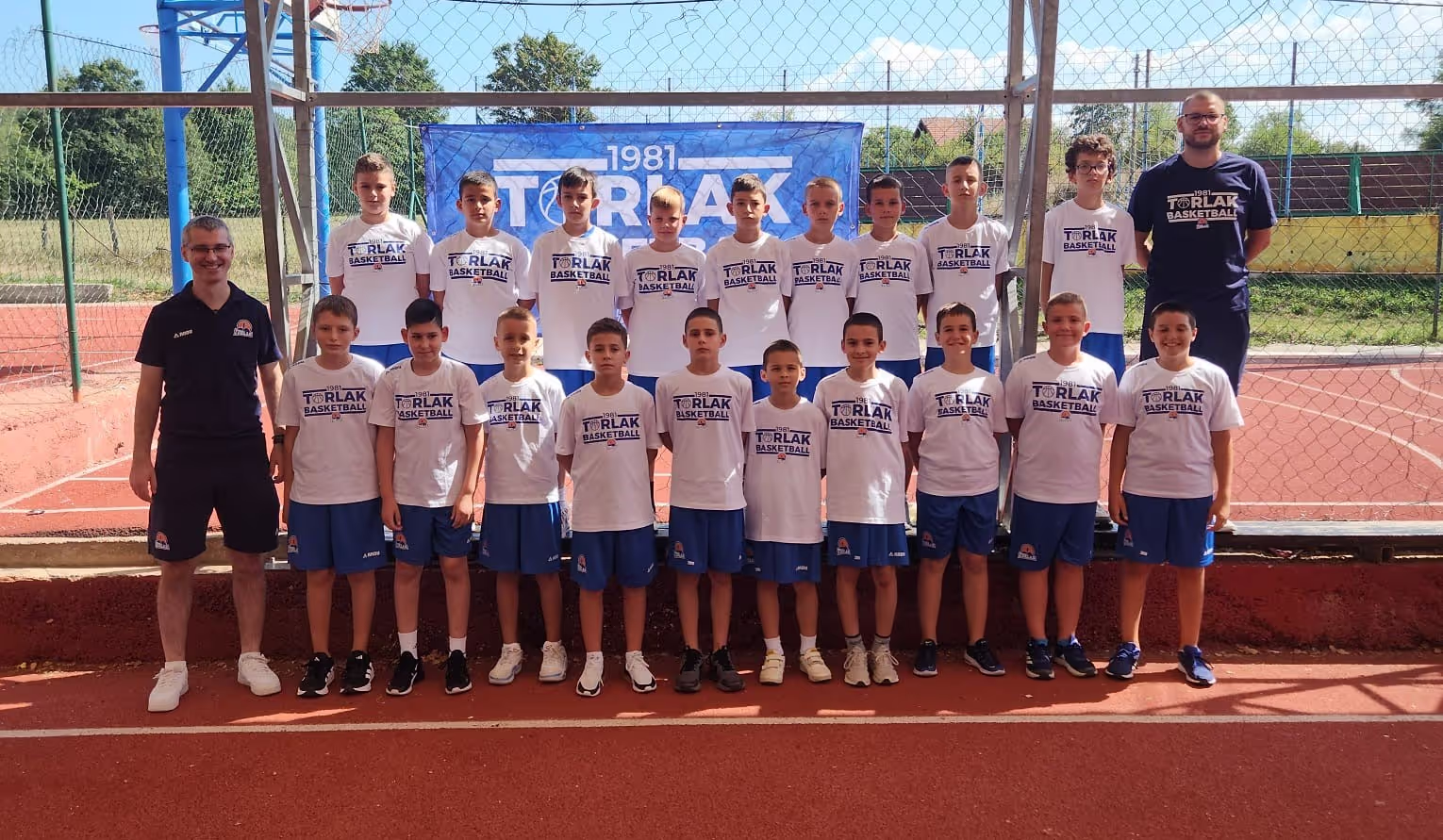 Youth basketball team posing in two rows on outdoor court wearing white Torlak Basketball shirts.