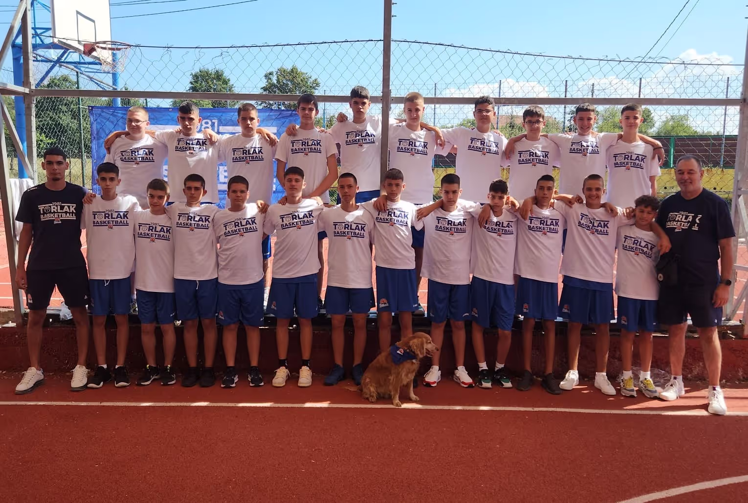 Youth basketball team wearing white T-shirts and blue shorts posing on outdoor court with a dog.