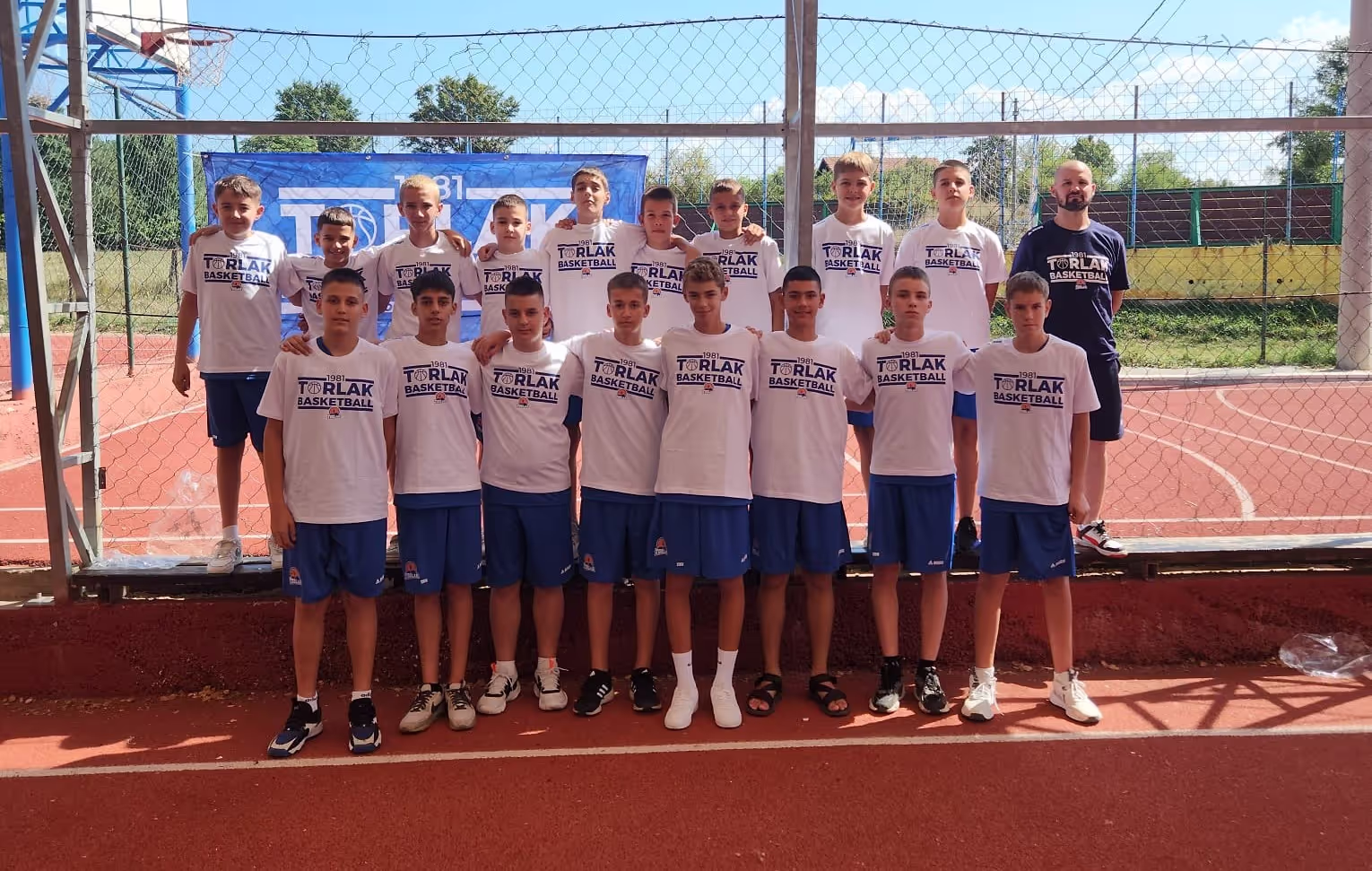 Youth basketball team of boys wearing matching white shirts and blue shorts posed in front of a fenced court.