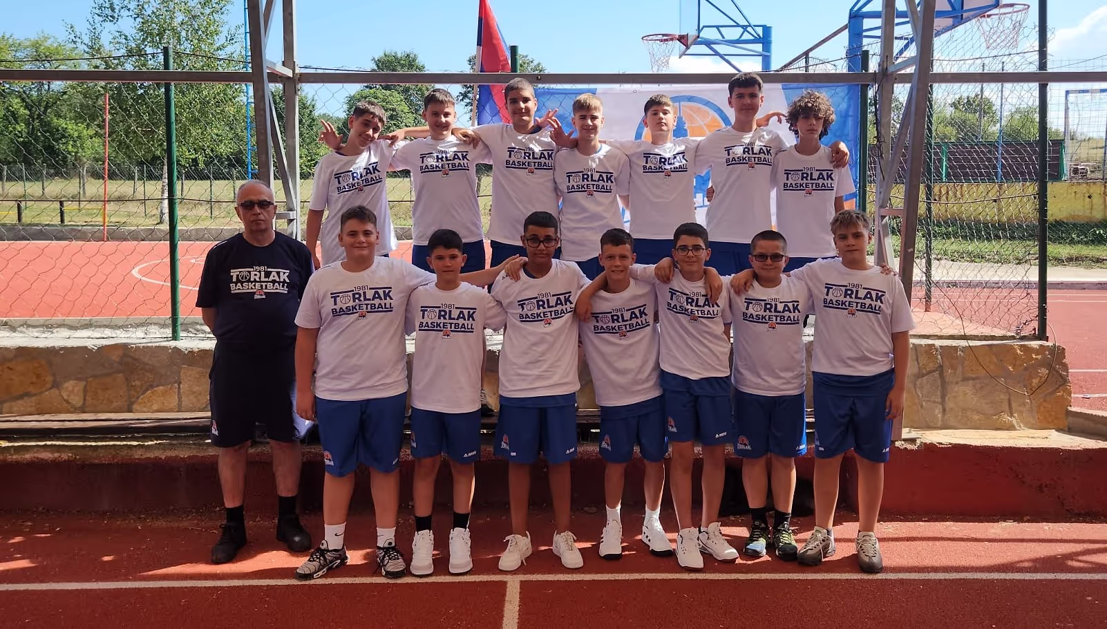 Youth basketball team in white shirts and blue shorts posing with coach on outdoor court.