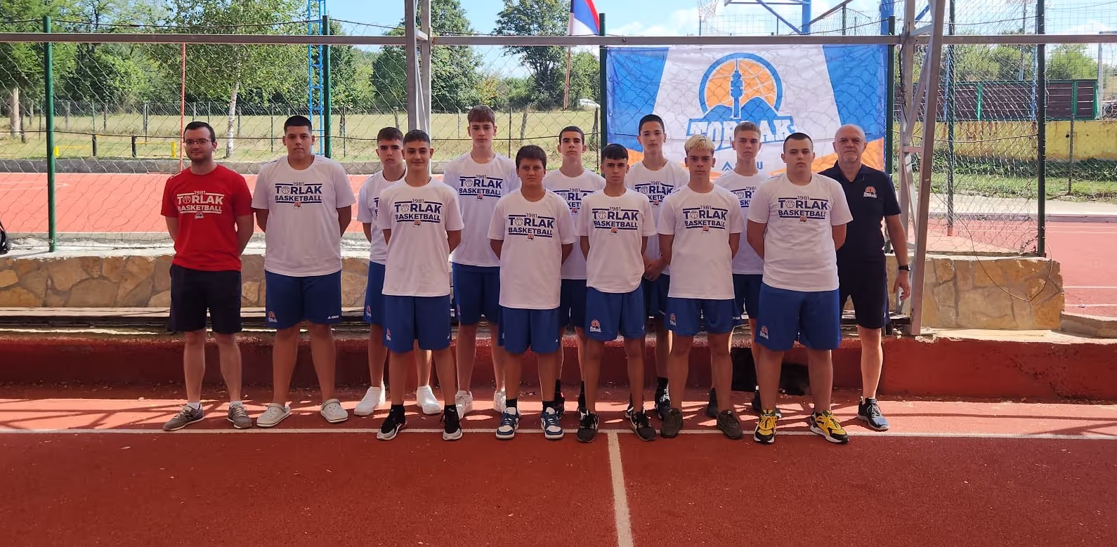 Youth basketball team posing on outdoor court in white T-shirts and blue shorts with coaches on each side.