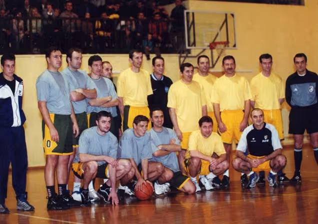Men's basketball teams posing on court, one in gray shirts, one in yellow shirts under gym hoop.