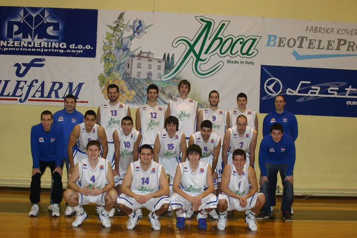 Basketball team of 15 men in white Aboca uniforms posing in indoor court with banners behind them.