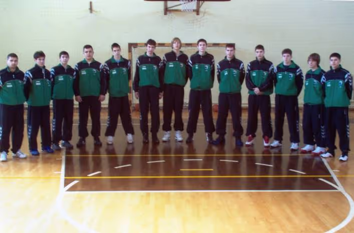 Eleven young men in matching green and black sportswear standing in a gym basketball court under a hoop.