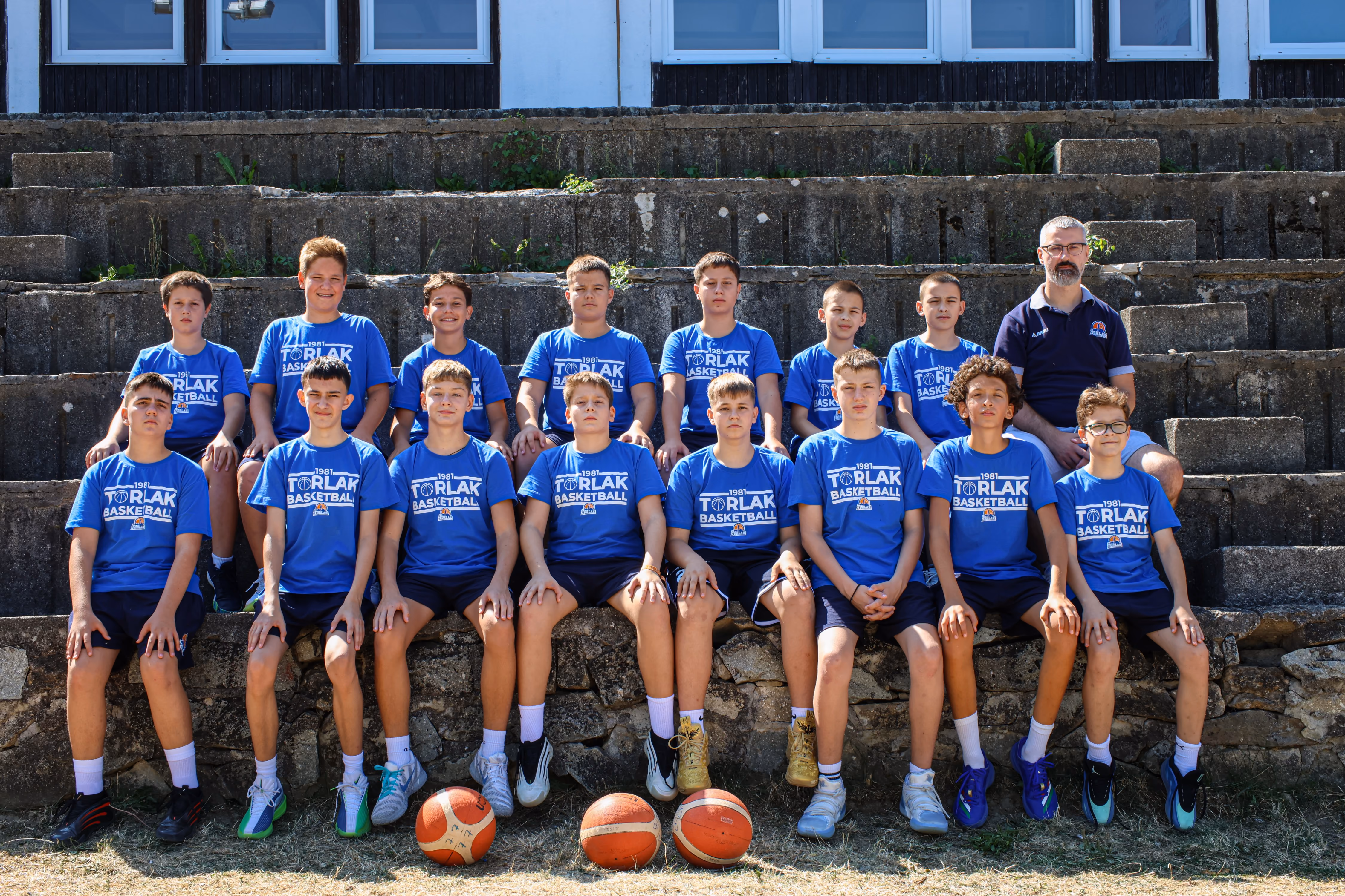 Youth basketball team of boys wearing matching white shirts and blue shorts posed in front of a fenced court.