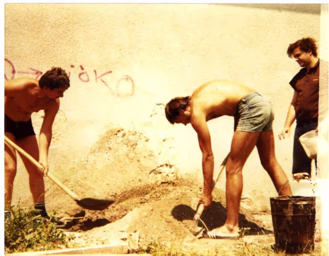 Three young men working outdoors with shovels and a bucket near a wall with graffiti.